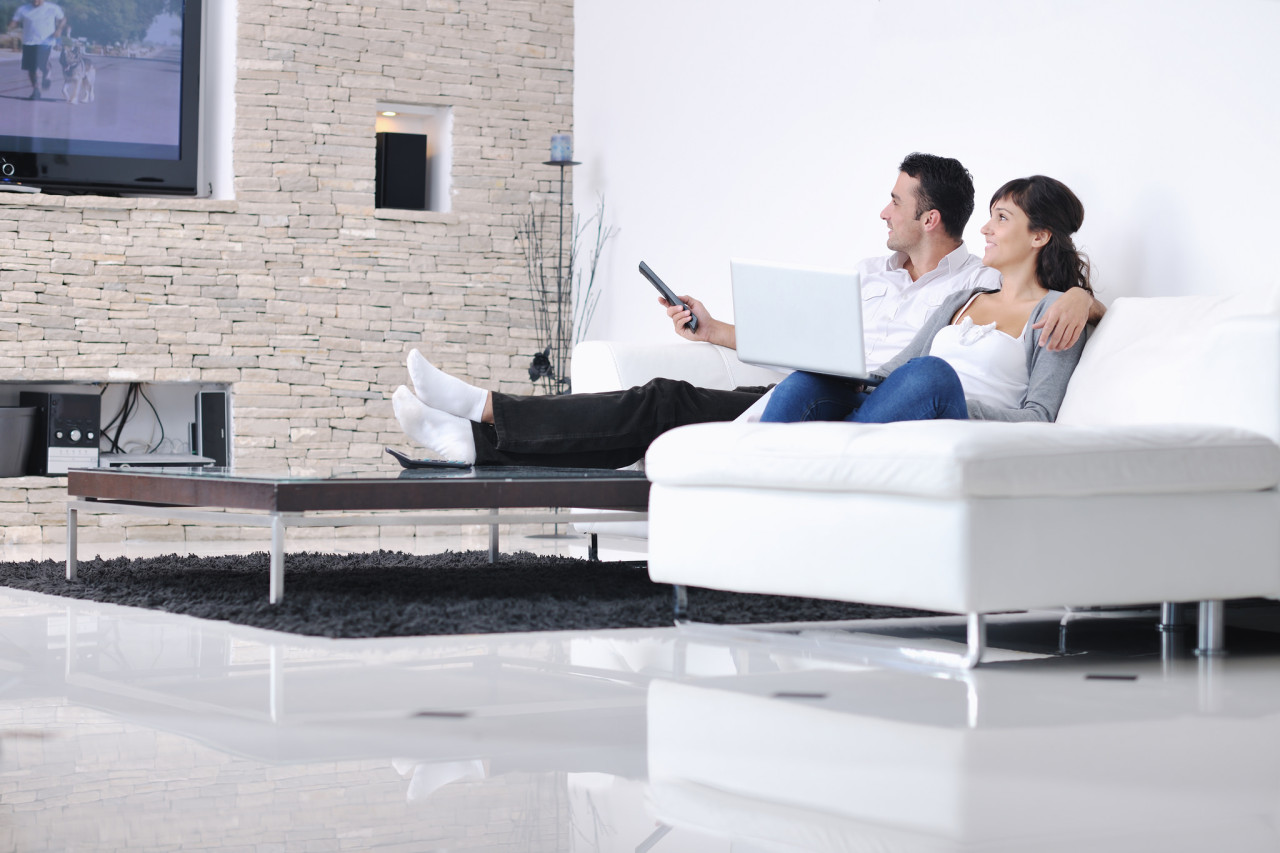 A couple lounges on a white sofa in a modern living room, watching TV and using a laptop.