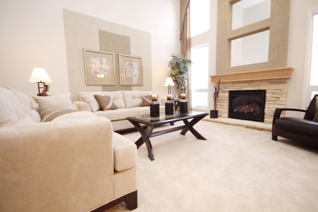 A spacious, well-lit living room with beige sofas, a glass-top coffee table, and a stone fireplace.